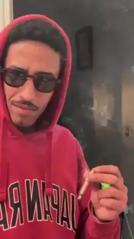 The video shows a man in a red hoodie and sunglasses, holding a lit cigarette. He is looking at the camera with a slight smirk. There is smoke in the air, and the background is a grey wall with a picture frame. The hoodie has the word "JAPAN" written on it.