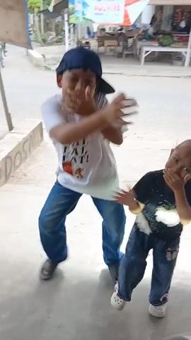 Two young children, a boy and a girl, are dancing in front of a building. The boy, wearing a blue baseball cap and a white t-shirt with an image of a strawberry and text, dances with his arms crossed and hands near his face. The younger girl, in a black t-shirt with a white graphic and blue jeans, mirrors some of his movements. They are standing on a paved surface, possibly a sidewalk, with a road and a building in the background. A motorcycle is visible in motion in the background. The scene appears to be outdoors during the daytime.