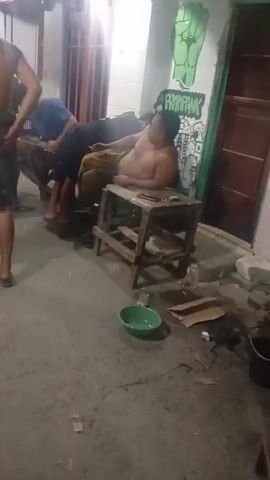 Two men are visible in this short video. One man is shirtless, wearing a yellow sarong, and is seated on a wooden stool. He is holding a cigarette. Another man, wearing a blue t-shirt, is standing and leaning over the seated man, seemingly interacting with him. They are in an outdoor environment, possibly an alley or courtyard, with a wall featuring graffiti behind them. The ground is paved and appears somewhat cluttered with debris. The lighting suggests it is either nighttime or a dimly lit area.