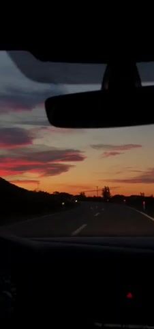 The video depicts a car driving along an asphalt road at sunset. The view is from inside the car, looking through the windshield, which is partially obscured by the dashboard.  The rear-view mirror is visible at the top of the frame, silhouetted against the colorful sky. The sky displays shades of orange, red, and pink, with patches of grey clouds. The road stretches into the distance, marked with a white line. There are trees and vegetation on either side of the road. Power lines are visible against the sky.  Music plays in the background.