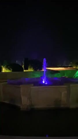 The video shows a fountain at night. The fountain is lit with blue and green lights. The water sprays up from the center of the fountain. The background is dark, with some trees and lights visible in the distance. The fountain is made of stone.