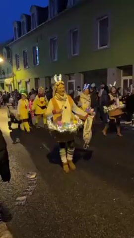A woman in an Easter-themed costume stands in the street, handing out candy. She wears bunny ears, a puffy vest, and leg warmers. She also sports a light-up skirt adorned with Easter decorations. Behind her, a parade of people in colorful costumes stretches down the street, which is lined with buildings. The event appears to be festive, and the woman seems to be enjoying her role in the celebration.