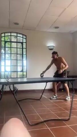 The video shows two people playing ping pong in an indoor room. The room has a brown tile floor and white walls with wall sconces.  There is a large window with black bars on the side of the room. A person with a black shirt and white hat stands on one side of the ping pong table, ready to hit the ball with a red paddle. On the other side of the table stands a shirtless person with dark patterned shorts.  The shirtless person hits the ping pong ball toward the person in the black shirt.
