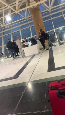 The video shows the interior of an airport terminal. The space has high ceilings with exposed steel beams and large windows. There are people standing near a check-in counter, and others are seated in waiting areas. A red suitcase is visible in the foreground. The floor is made of marble tiles with black and green geometric designs. There is a digital screen showing a nature scene. The camera moves slightly, causing blurring.