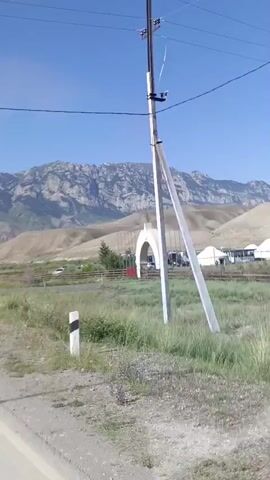 A bright, clear day with a blue sky dominates the video. In the foreground, a dirt road is visible. A large white archway stands on the left side of the frame, with a wooden fence in front of it. Beyond the fence, several white tents or yurts are set up, along with a building. Various cars are parked in front of the building and tents. In the background, rolling hills with sparse vegetation lead up to a majestic mountain range. A utility pole stands in the right foreground of the image. The camera appears to be moving, suggesting the video was shot from a vehicle.