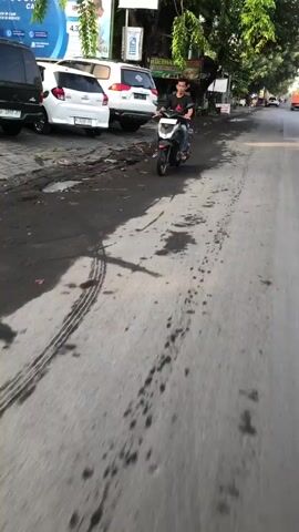 A young male rides a scooter down a paved road. He is wearing a dark shirt and light-colored pants. The road is wet in patches, and there are tire marks visible. In the background, several cars are parked on the side of the road, and there are trees and buildings. The sun is shining, casting shadows on the road.