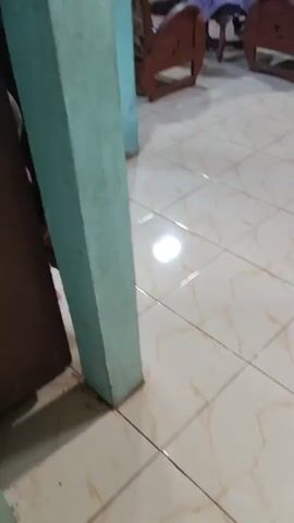 The video shows a floor with light-colored tiles. A light green-painted pillar is visible on the left side of the frame. In the background, a cardboard box is partially visible. The floor appears to be clean with some reflections from lighting. There are no people visible in the video. The camera is moving, providing a panning shot of the floor and a portion of the wall.