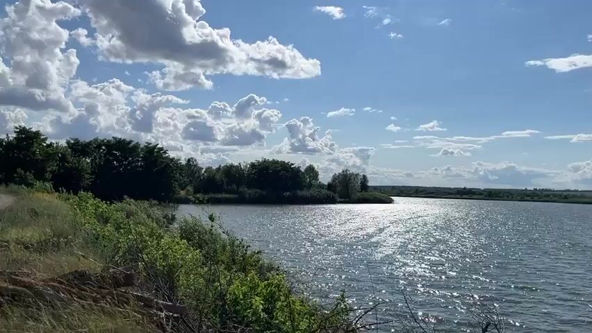 The video opens with a view of a lake on a sunny day with scattered clouds. A dirt road runs along the grassy bank to the left, bordered by bushes and trees. The water is calm and reflects the sunlight. As the camera pans, a man wearing a black baseball cap and sunglasses is seen from behind, looking out at the lake. The camera continues to pan to the right, revealing two more men standing on a wooden platform near the water. One man, wearing a blue shirt and black shorts, is facing the camera and gesturing with his hand. The other man, wearing a white t-shirt and dark shorts, is facing away from the camera. The sky is blue with dramatic white clouds.