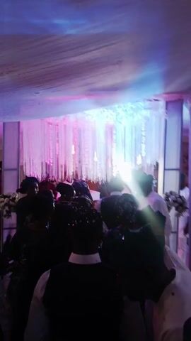 The video depicts a gathering, likely a celebration of some sort, taking place in a decorated indoor space. The initial shots focus on a crowd of people dancing, with decorations including draped white material, string lights in pink and green hues, and floral arrangements visible in the background. The video then pans to show a seated group of people at tables adorned with floral centerpieces, decorative plates, and what appear to be refreshments. Some people are dressed in traditional attire. There are some speakers visible in the background and windows. The lighting conditions are somewhat dim, adding to the festive atmosphere.