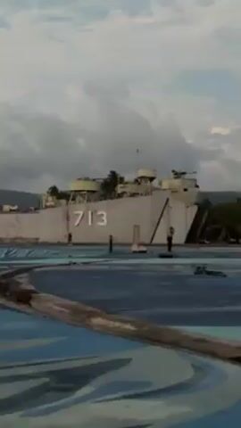 The video shows a beached U.S. Navy tank landing ship, specifically identified by the hull number 713, adjacent to a blue and white painted concrete area reminiscent of a drained swimming pool or decorative area.  A few people are seen near the ship. The environment appears to be an outdoor area with mountains visible in the background under an overcast sky. The video concludes with a view of the scene from what appears to be a low vantage point with objects, including a plastic bag, visible on the foreground surface.