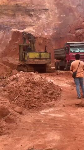 The video shows a construction site with red dirt. An excavator is loading dirt into a dump truck. A man is standing near the dump truck, and another person is partially visible on the right. A white container with a red lid is on the ground near the man. The excavator is yellow and has tracks. The dump truck is red and appears to be a commercial vehicle. The terrain is uneven and appears to be a mine or quarry. The overall scene depicts active construction or excavation work.