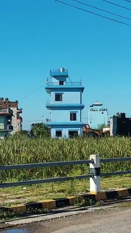 A three-story blue building with a water tank on top is visible behind a tall grassy field. The building has multiple windows. A smaller blue building is further in the background. The sky is clear and blue.