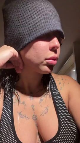 A woman with a gray beanie hat and a black and white polka dot sports bra is seen in a close-up shot. She has tattoos on her chest and shoulder. She adjusts the beanie, pulling it back from her face. The background is slightly out of focus. Music is playing in the background.