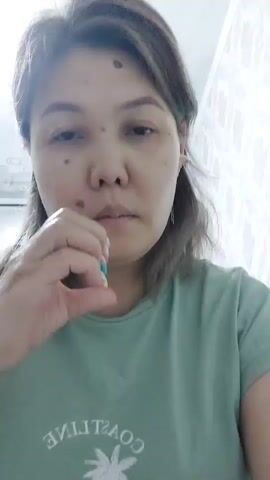 A woman with brown hair and a mole on her forehead makes a heart shape with her hands in front of her mouth. She is wearing a green t-shirt with a graphic design and a gold ring on her left ring finger. The background appears to be an indoor setting with a textured wall.