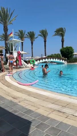 A group of people, mostly children, are swimming and playing in a large outdoor swimming pool. There are several palm trees surrounding the pool, and lounge chairs and umbrellas are visible in the background. The sky is clear and blue. One person is standing on a platform near a slide, and another is on a lifeguard stand. There are several inflatable pool toys in the water, including noodles and rings. The poolside is paved with tiles.