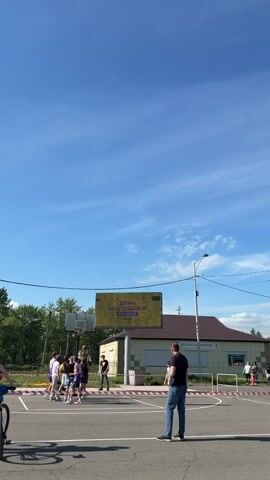 A group of young people are playing basketball on an outdoor court on a sunny day with a clear blue sky and scattered clouds. A large yellow billboard with Russian text hangs above the basketball hoop, reading "ДЕНЬ МОЛОДЕЖИ ЕЛИЗОВО" (Youth Day Yelizovo). A man in a black t-shirt and blue jeans stands watching the game. In the background, there is a building with the text "ДЕТСКАЯ БИБЛИОТЕКА" (Children's Library) visible. The scene appears to be a community event.