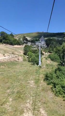 The video shows a POV shot from a ski lift ascending a grassy mountainside on a clear sunny day. The ski lift towers and cables are visible, as are the empty chairs of the lift. The surrounding landscape is covered in green grass and scattered trees, with a dirt path visible on the left side of the frame. The top of the mountain is visible in the distance, with more ski lift infrastructure. The video is approximately 4 seconds long.