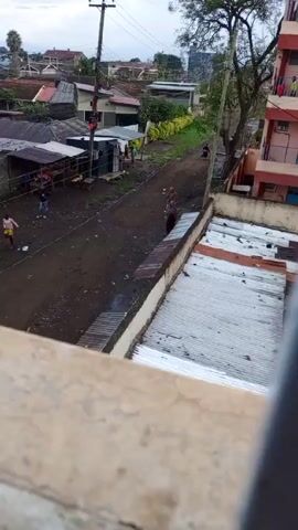 The video shows an overview of a neighborhood street in what appears to be a rural or urban-fringe area. The street is unpaved and appears muddy or dusty, with a few pedestrians visible. One individual walks down the street wearing a brown head covering and a patterned dress. Other people can be seen standing around what looks like a small shack or store. Overhead power lines are visible along with utility poles. The roofs of the surrounding buildings are made of corrugated metal sheets, some showing signs of rust. The video seems to be taken from an elevated position, possibly from a building overlooking the street.