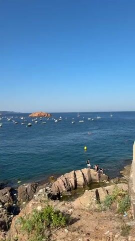 This video captures a lively summer scene at a beach in Tossa de Mar, Spain. The bright, clear blue sky indicates a sunny day, likely mid-morning or early afternoon. The turquoise water of the Mediterranean Sea is calm, dotted with numerous boats of various sizes anchored or gently sailing.

On the rocky shoreline in the foreground, several people are enjoying the warm weather. Some are seated on the rocks, while others are swimming in the clear water. Yellow buoys are visible in the sea, possibly marking swimming areas or boat moorings.

Further out, a picturesque bay unfolds. A crescent-shaped sandy beach is crowded with sunbathers, umbrellas, and beachgoers enjoying the water. Buildings, including hotels and houses, are nestled into the lush green hills that surround the bay, characteristic of the Costa Brava coastline. In the distance, another rocky island rises from the sea, adding to the scenic beauty of the location. The overall atmosphere is one of relaxation and summer vacation enjoyment, with people soaking up the sun and the refreshing sea. There is no visible text in the video.