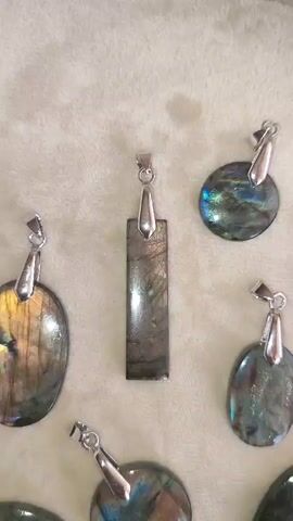 A close-up shot shows a collection of labradorite pendants displayed on a textured, light-colored surface. The pendants vary in shape and size, with several oval, round, and rectangular pieces visible. Each pendant is made of labradorite, a gemstone known for its iridescent, multicolored sheen, which displays flashes of blue, green, gold, and purple. The pendants are adorned with silver-toned bails, suggesting they are ready to be worn as jewelry. The lighting highlights the natural beauty and unique patterns of the labradorite, making the stones appear to shimmer and glow. The background is soft and out of focus, emphasizing the pendants as the main subject.