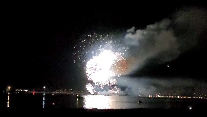 This is a nighttime video of a fireworks display over a body of water. Several large fireworks are exploding in the sky, creating bright flashes of white and yellow light. Smoke from the explosions drifts across the dark sky. In the foreground, the water reflects the light from the fireworks, and a dark silhouette of land with scattered lights can be seen in the distance. A few small boats are visible on the water.
