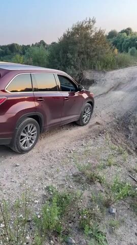 In Drakino, Russia, a maroon Toyota Highlander SUV navigates a dirt path with uneven terrain. The vehicle is positioned on the left side of the frame, its wheels partially on a raised section of the path and partially in a rut. Sunlight reflects off the windows of the SUV, suggesting it's either early morning or late afternoon.

The scene is an outdoor setting with dry vegetation and some green bushes and trees in the background. A person stands further up the path on the right, observing the vehicle. The sky above is clear with a gradient of colors indicating either sunrise or sunset. The ground is dusty and rocky, characteristic of a rural or unpaved road.

The activity appears to be off-road driving, with the SUV possibly maneuvering through a challenging section of the path. The focus is on the vehicle and its interaction with the terrain.

Visible text on the license plate of the SUV reads "U97BP 777". The "V6" badge is also visible on the rear of the vehicle. The overall impression is one of a leisurely drive in a natural, somewhat uncultivated environment. The time of day creates a peaceful atmosphere.