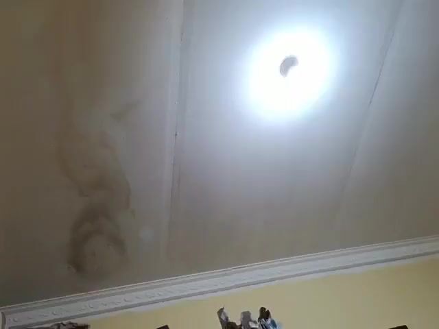 A close-up video shows a stained ceiling with a light fixture. The stains are discolored and appear to be water damage. The ceiling is white and has a textured finish. The light fixture is round and has a bright bulb. The video pans slightly across the ceiling, showing the extent of the discoloration. The overall impression is one of a neglected or damaged ceiling.