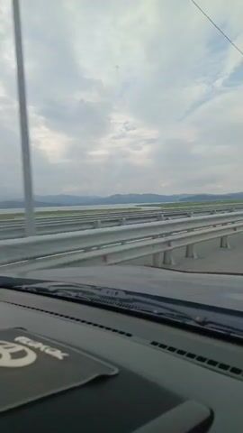 The video captures a view from inside a vehicle driving along a highway. The perspective is from the dashboard looking forward through the windshield. The highway has multiple lanes separated by metal guardrails. In the distance, beyond the highway, is a body of water with hills or mountains on the horizon. The sky is cloudy. Other vehicles can be seen on the highway in the distance. A windshield wiper is visible in the foreground.