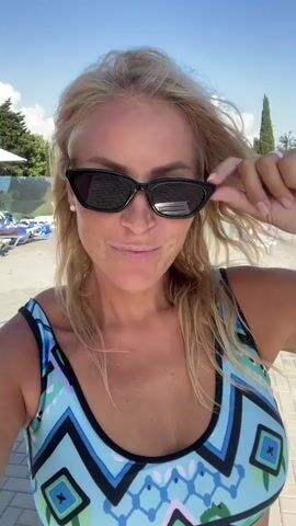A blonde woman with sunglasses on top of her head smiles at the camera. She is wearing a blue and white patterned bikini. The background shows a bright, sunny day with trees and a pool area. She is adjusting her sunglasses with her right hand. The video is a selfie style shot.