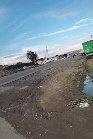 The video pans from a dusty roadside to a paved road. On the left, there is a dirt path with tire tracks and some puddles. In the distance, there are several white vans and cars parked. Further back, there are buildings and trees. A tall, thin structure, possibly a radio tower, is visible. A large green truck with white lettering is on the right side of the paved road. The sky is blue with some white clouds. The camera continues to pan to the right, showing more of the paved road and the landscape beyond. On the right side of the road, there is more vegetation and a small puddle with some debris. The scene appears to be a rural or semi-urban area with ongoing construction or development, suggested by the unfinished buildings on the hills in the background. The lighting suggests it might be late afternoon or early morning.