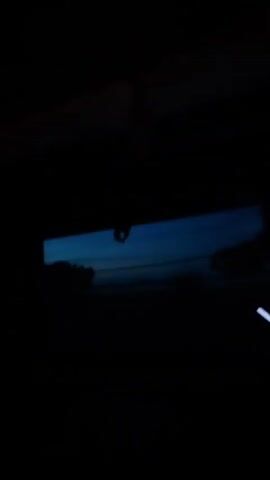 This is a dark, shaky video shot from inside a vehicle. The camera is pointed towards the rearview mirror and the windshield. The sky outside is a deep blue, suggesting it is either dawn or dusk. The silhouette of trees or other foliage can be seen against the sky. The interior of the car is mostly black, but there are a few faint lights visible, possibly from the dashboard. The video is very short, only a few seconds long.