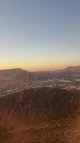 From a window seat on an airplane, a view of the sun setting over mountains and the Mediterranean Sea is visible. The wing of the plane appears in the frame. The plane appears to be descending toward an airport.