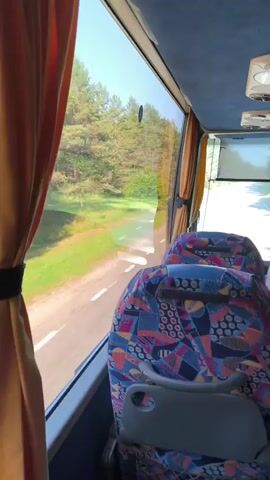 The video shows the interior of a moving bus, looking out the window. The window is partially covered by an orange curtain. Outside the window, a road can be seen curving through a forest. The trees are tall and green, and the sky is blue with some clouds. The road has white lines marking the lanes. Inside the bus, several seats are visible. The seats have a colorful, geometric pattern. The camera is positioned to look out the window, with the seats in the foreground. The movement of the bus creates a blurred effect on the landscape outside.