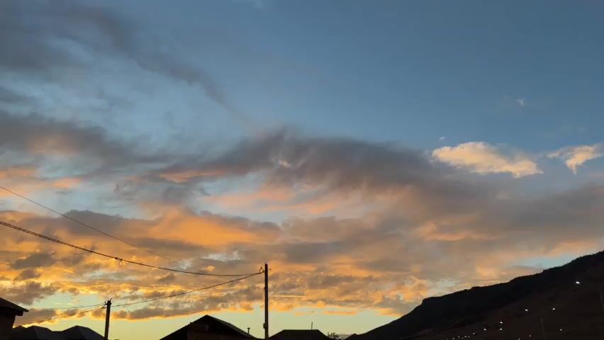 The video displays a wide shot of a rural mountainous landscape at dusk. The sky is a mix of clear blue with scattered clouds tinged with orange and yellow hues, indicative of a sunset. The foreground shows a few houses with dark roofs, power lines stretching across the frame, and a dirt road. In the mid-ground, a road is visible with a few cars and trucks driving on it, bordered by a fence. The background is dominated by a large, dark mountain range, sparsely vegetated with a few scattered lights visible along its base, suggesting a settlement or roadside illumination. A streetlamp stands prominently on the right side of the frame. The overall atmosphere is peaceful and serene.