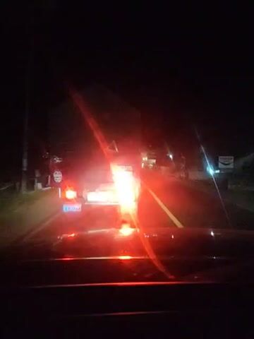 The video shows a first-person perspective from inside a vehicle driving at night. The headlights of the vehicle illuminate a road ahead, revealing a truck in front with its brake lights illuminated. Other vehicles and roadside lights are also visible in the distance. The road appears to be asphalt, and there are some structures and vegetation along the sides. The overall scene suggests a journey on a rural road after dark.