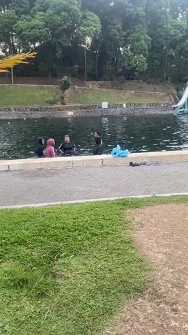 A group of people are in the water at a pool. There is a slide in the background. The people are wearing swimwear. One person is wearing a pink headscarf and a dark swimsuit. Another person is wearing a black headscarf and a dark swimsuit. Another person is wearing a black headscarf and a dark swimsuit. Another person is wearing a black headscarf and a dark swimsuit. A person is standing on the edge of the pool wearing a black swimsuit. There are several blue floatation devices in the water. There are trees in the background. There is a grassy area in the foreground. The sky is overcast.