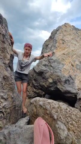 A woman wearing a white t-shirt, grey shorts, and a pink baseball cap is climbing on rocks. She is barefoot and has braids. A pink backpack is visible in the foreground. The environment appears to be a rocky coastline with the ocean in the background and a cloudy sky.