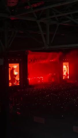 A wide shot of a concert venue. The stage is lit with red and yellow lights, with two large screens on either side displaying vibrant imagery. A large crowd of people is visible in front of the stage, many holding up illuminated phones. The overall atmosphere is energetic and dynamic.