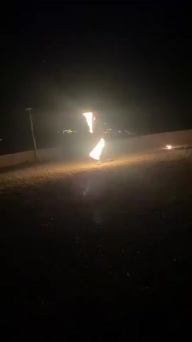A person is juggling two flaming torches in a dark outdoor setting. The torches are lit and appear to be creating bright streaks of light as they are spun. The person is wearing dark clothing and is standing in an open area that appears to be a grassy or dirt patch. In the background, there is a hint of distant lights, suggesting a town or city. The overall scene is illuminated by the flames of the torches and a faint glow from the distant lights.