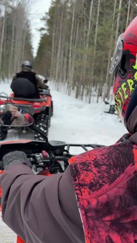 Here's a description of the video:

The video shows a first-person perspective of someone riding an orange ATV on a snow-covered path through a forest. The rider wears a helmet with red designs and a brown jacket. Another ATV, also orange, is ahead on the trail, driven by someone wearing a black helmet and a dark jacket. The side mirror of the first ATV reflects the driver holding a phone, presumably taking a picture or video. The trees on either side of the path are mostly bare. The overall scene is one of a winter ATV ride through a wooded area.