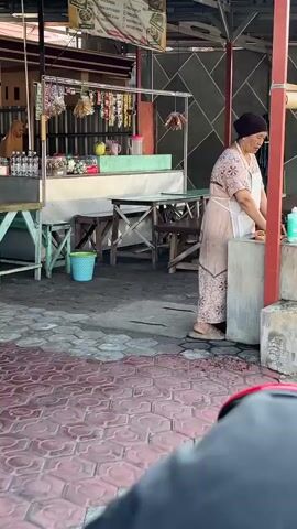 The video shows a woman wearing a light-colored patterned dress and a white apron standing at a counter in what appears to be an outdoor food stall. She has a dark headscarf on. In front of her, there is a gray concrete structure, possibly a sink or preparation area, and a small white container. To her left, there is a wooden table with a yellow bucket underneath it. Behind her, there are stalls with various items for sale, including what look like food packets hanging. A dog is visible sitting near one of the stalls. The ground is paved with red and gray interlocking tiles. The scene is outdoors with a roof structure overhead, indicated by the presence of pillars and lighting. The overall impression is that of a busy marketplace or food court.