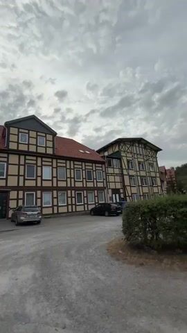 The video shows a walk-through of a street in front of several buildings. The buildings appear to be residential or perhaps small businesses, with a mix of architectural styles. Some buildings have a half-timbered facade, while others are more modern. Cars are parked in front of some of the buildings. The weather appears to be overcast. The camera pans and moves, showing different angles of the street and buildings. There are no people visible in the video.