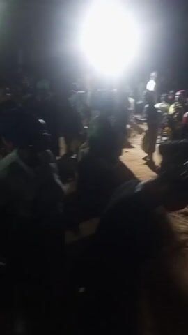 The video depicts a nighttime gathering of people in an outdoor setting, illuminated by a bright, possibly artificial, light source. The majority of the individuals are dressed in casual clothing and are standing or moving around. The ground appears to be dirt or sand, suggesting a rural or less developed area.  Many of the people are wearing head wraps. There's a sense of activity and movement, but the exact nature of the gathering is not clear from the visual information.