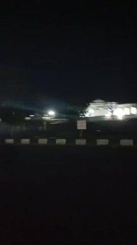 The video shows a nighttime drive past a modern building and a row of flags. The building is white with bright lights. The flags are on tall poles and are illuminated. The road has white and black markings. There are some trees and lights visible in the background. The video is shaky, indicating it was filmed from a moving vehicle.
