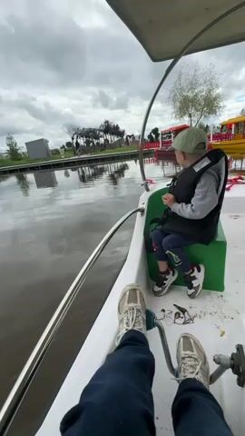 The video shows a first-person perspective from inside a small pedal boat. In the foreground, a person's legs and feet wearing blue jeans and white sneakers are visible, with their feet positioned near the boat's pedals. To the right and slightly behind, a young boy wearing a light green cap, a gray hoodie over a dark vest, and dark pants with colorful stripes on the legs sits facing away from the camera. He is seated on a green plastic box. The boy is looking out towards the water and the distant shore. The boat is on a body of water that appears calm, with reflections of the cloudy sky and distant trees visible on its surface. In the background, there is a shoreline with wooden structures, some trees with sparse foliage, and other colorful pedal boats docked or in the water. The sky is overcast with gray clouds.