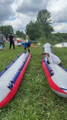 A group of people are preparing inflatable boats for use near a body of water. Two fully inflated boats, red, blue, and gray, lie on the grass. One person is bent over one of the boats, adjusting something.  Another person is half inside the other boat. Other people are standing or walking around the boats in the background. In the distance, there is a calm lake or river, and the sky is mostly cloudy.