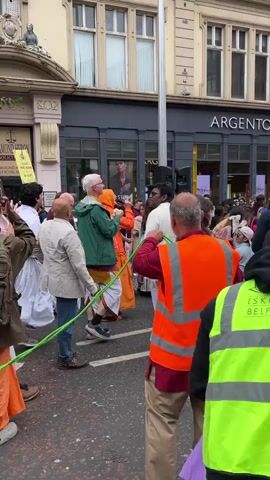 A group of people are gathered in a street with buildings on either side. Some individuals are wearing traditional Indian clothing. A person in an orange safety vest is holding a rope that is being pulled by others. There appears to be a procession or celebration taking place. The time stamp is 00:00 to 00:05.