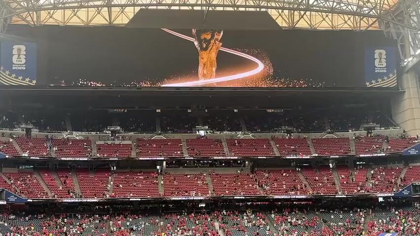 This media file depicts a wide-angle view of a crowded stadium, likely a sports arena, from the perspective of someone seated in the stands. The primary focus is on a massive Jumbotron screen suspended above the field. The screen displays a dynamic graphic featuring the FIFA World Cup trophy, surrounded by a swirling, glowing ribbon of light and sparkling particles. On either side of the trophy graphic are banners indicating "FIFA WORLD CUP 2026".

Below the Jumbotron, rows upon rows of red stadium seating are visible, filled with a large number of spectators. Many of the people in the stands are wearing red clothing, suggesting they are supporting a particular team or participating in a themed event. The stadium appears to be enclosed by a roof structure with exposed metal beams and lighting fixtures.

The overall scene suggests an event or announcement related to the FIFA World Cup 2026, possibly a promotional ceremony or a kickoff event. The lighting on the screen is bright and dramatic, while the stadium itself is illuminated, suggesting it is either daytime or the stadium lights are on for an evening event. The atmosphere seems energetic and anticipatory, given the large crowd and the prominent display of the World Cup branding. The location is identified as Houston, United States of America, placing the event within the context of one of the host cities for the 2026 World Cup. There are no overt indications of weather conditions.