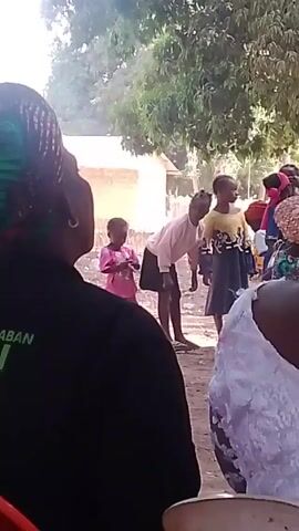 The video shows a group of people, including children and adults, gathered outdoors, possibly for a social event. The children are standing in front of the crowd, and one is actively dancing or moving to music. The adults are watching and seem to be enjoying the performance. The setting appears to be a village or rural area, with simple buildings and trees in the background. The atmosphere is lively and festive.