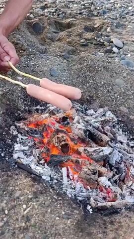 A close-up shot shows two sausages on skewers being held over a small campfire on a sandy beach. The fire consists of glowing embers and burning wood. The sausages are pale pink and appear to be raw. A person's hands are visible at the top of the frame holding the skewers. The background is blurred but shows sand and small pebbles.